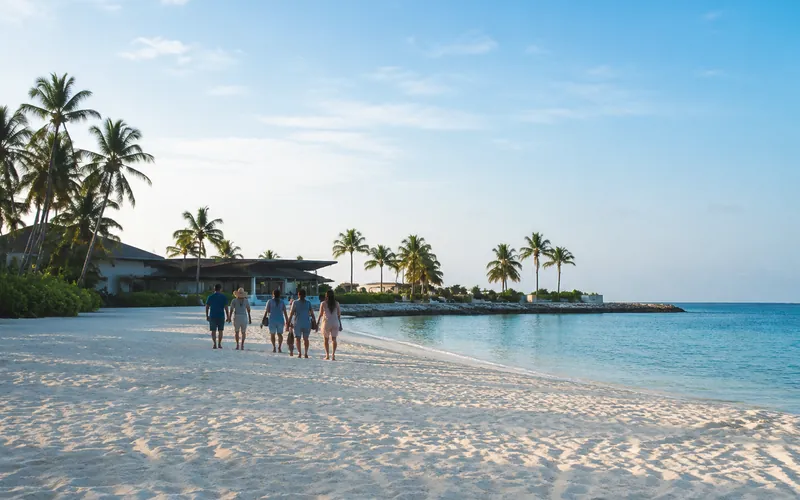 A group of travelers walking side by side along a wide, quiet stretch of pale sand beside calm turquoise water.