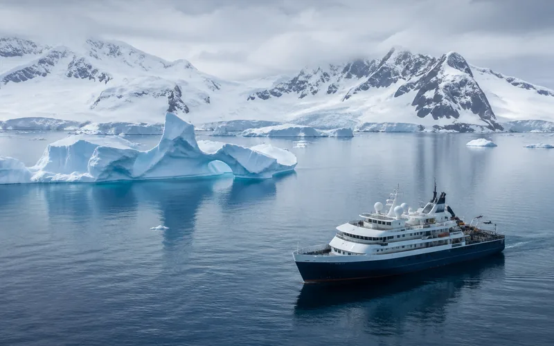 A small dark-hulled expedition ship in still polar water beside a sculpted blue iceberg, with snow-covered mountains under heavy cloud behind.