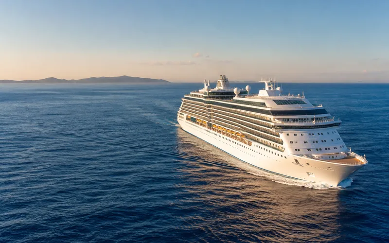 A large white multi-deck ocean cruise ship under way on deep blue water in late afternoon light, with a distant island ridge behind it.
