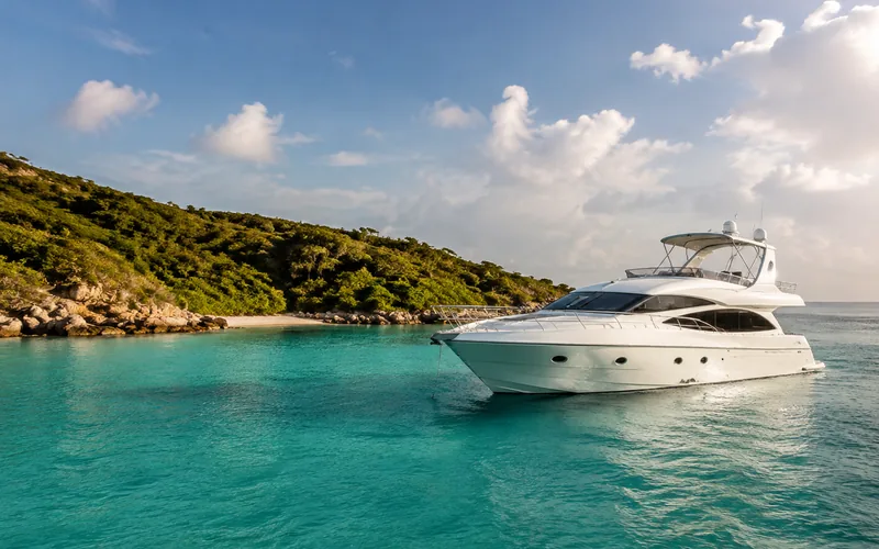 A white motor yacht at anchor in clear turquoise water beside a green headland and a small sandy cove under bright cloud.